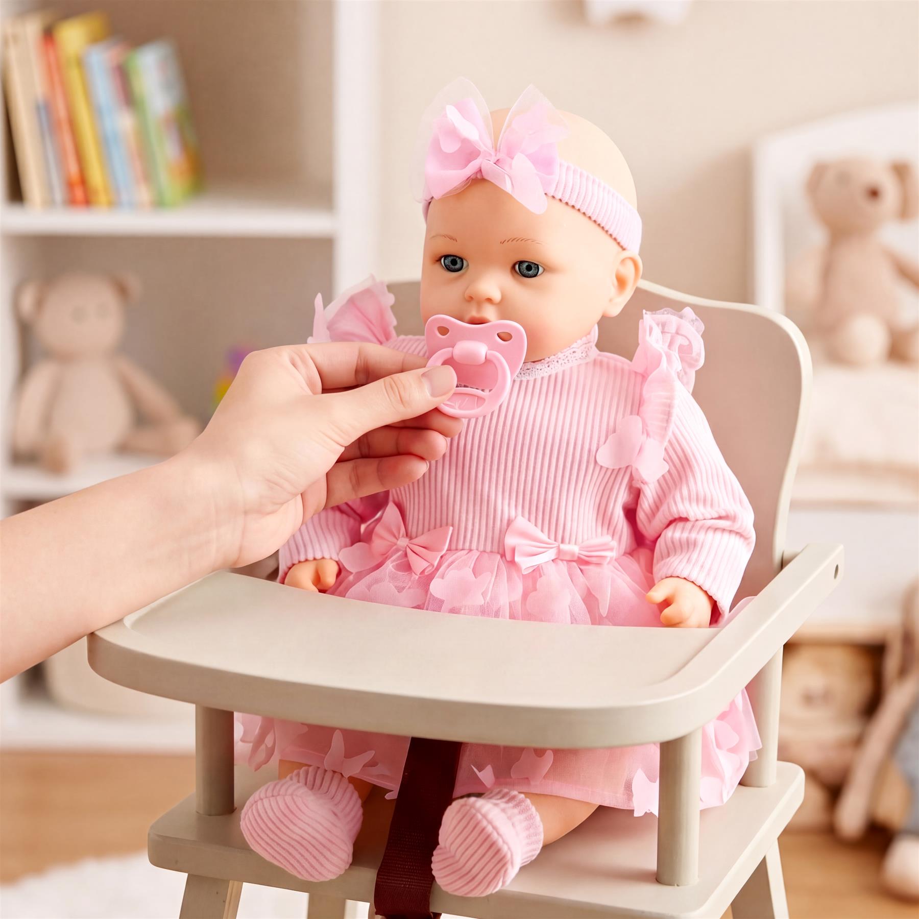 A hand holds a pink pacifier near a doll seated in a light-colored chair, wearing a pink outfit.