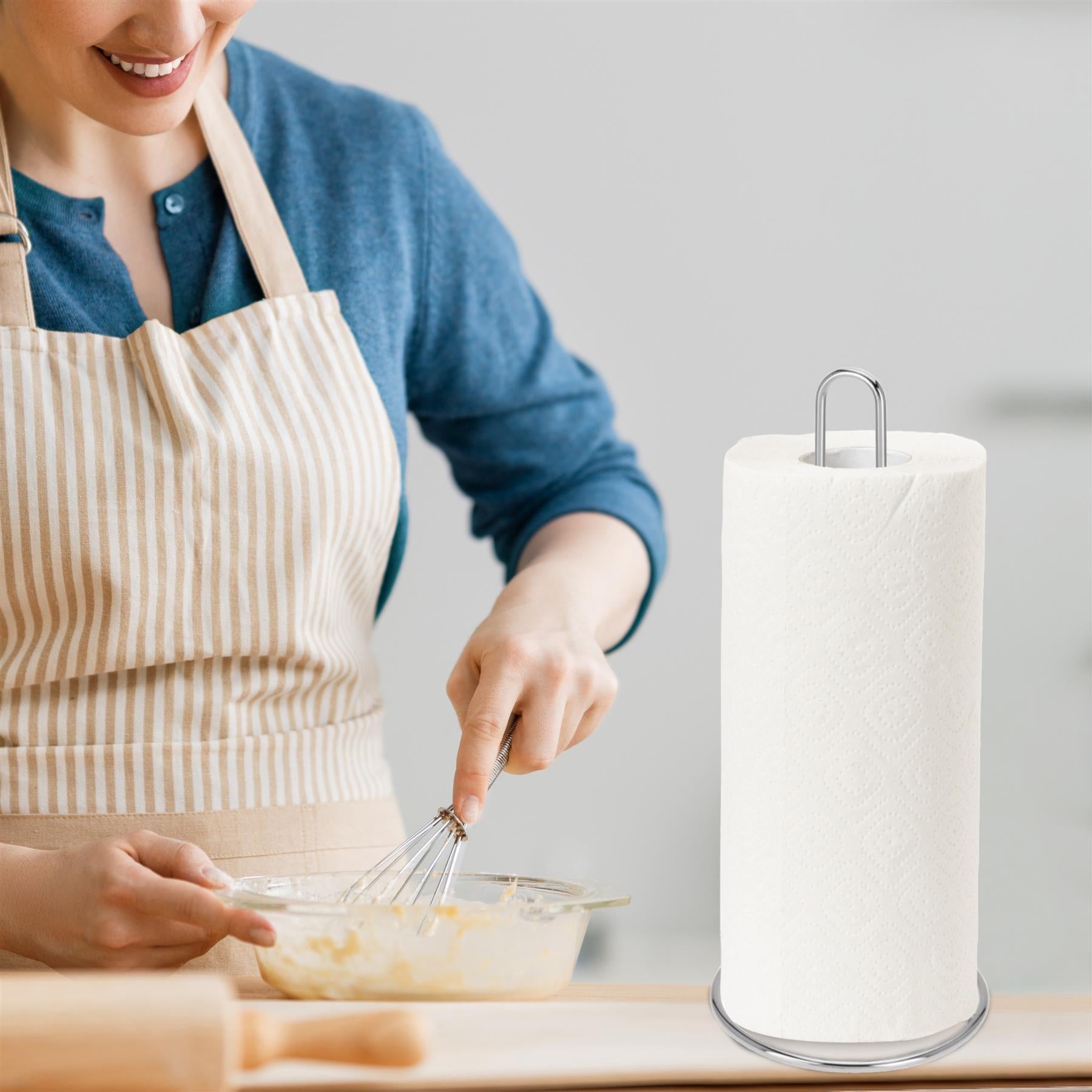 A woman in a kitchen, wearing a blue shirt and a beige and white striped apron, holding a bowl of yellow liquid and a whisk. Next to her is a paper towel roll with a metal base and a paper towel holder on top. - The Magic Toy Shop