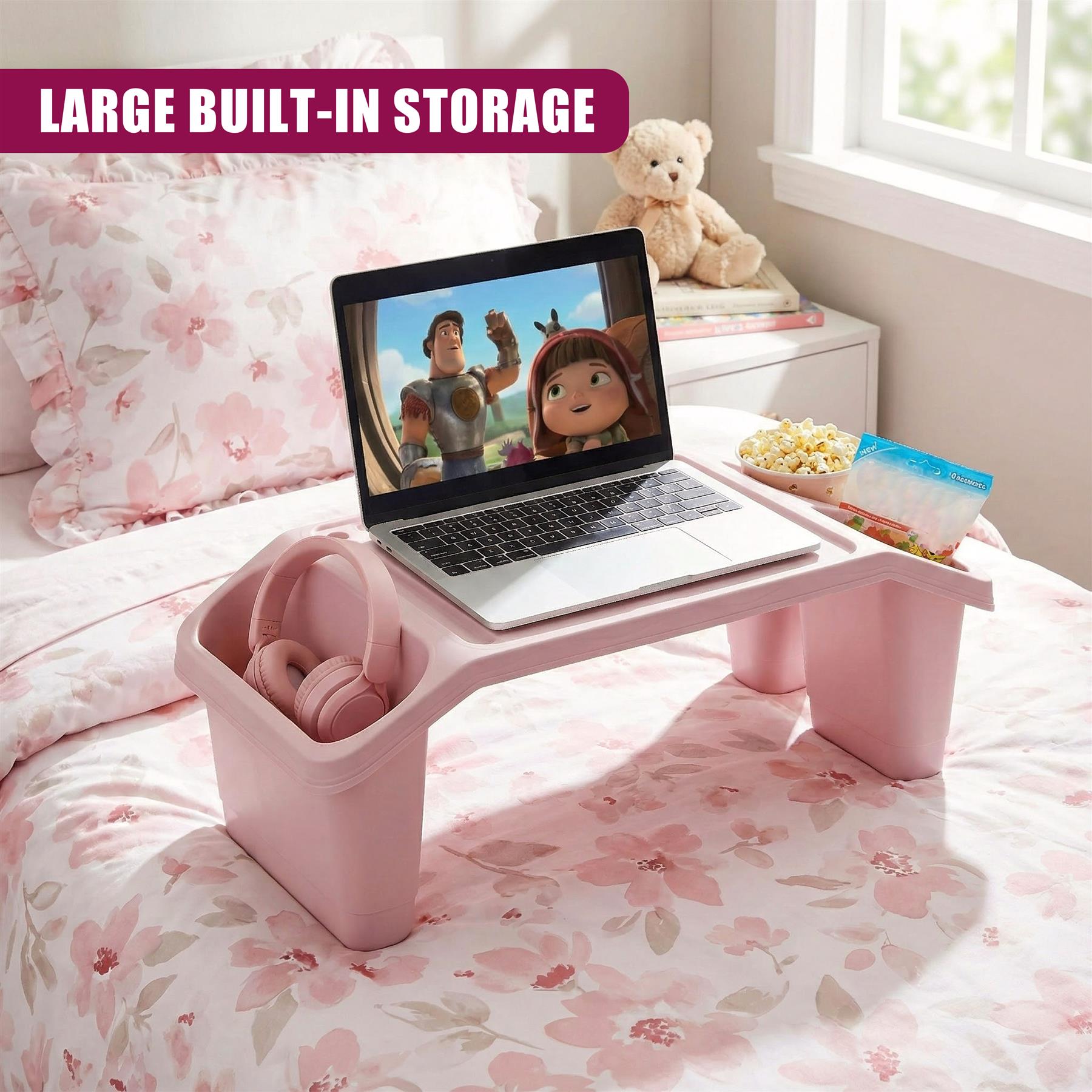 A laptop sits on a pink desk, displaying a cartoon scene. The desk has a built-in tray for snacks and a storage bin for headphones. It's placed on a bed with a pink floral duvet and a white wall with a window. - The Magic Toy Shop