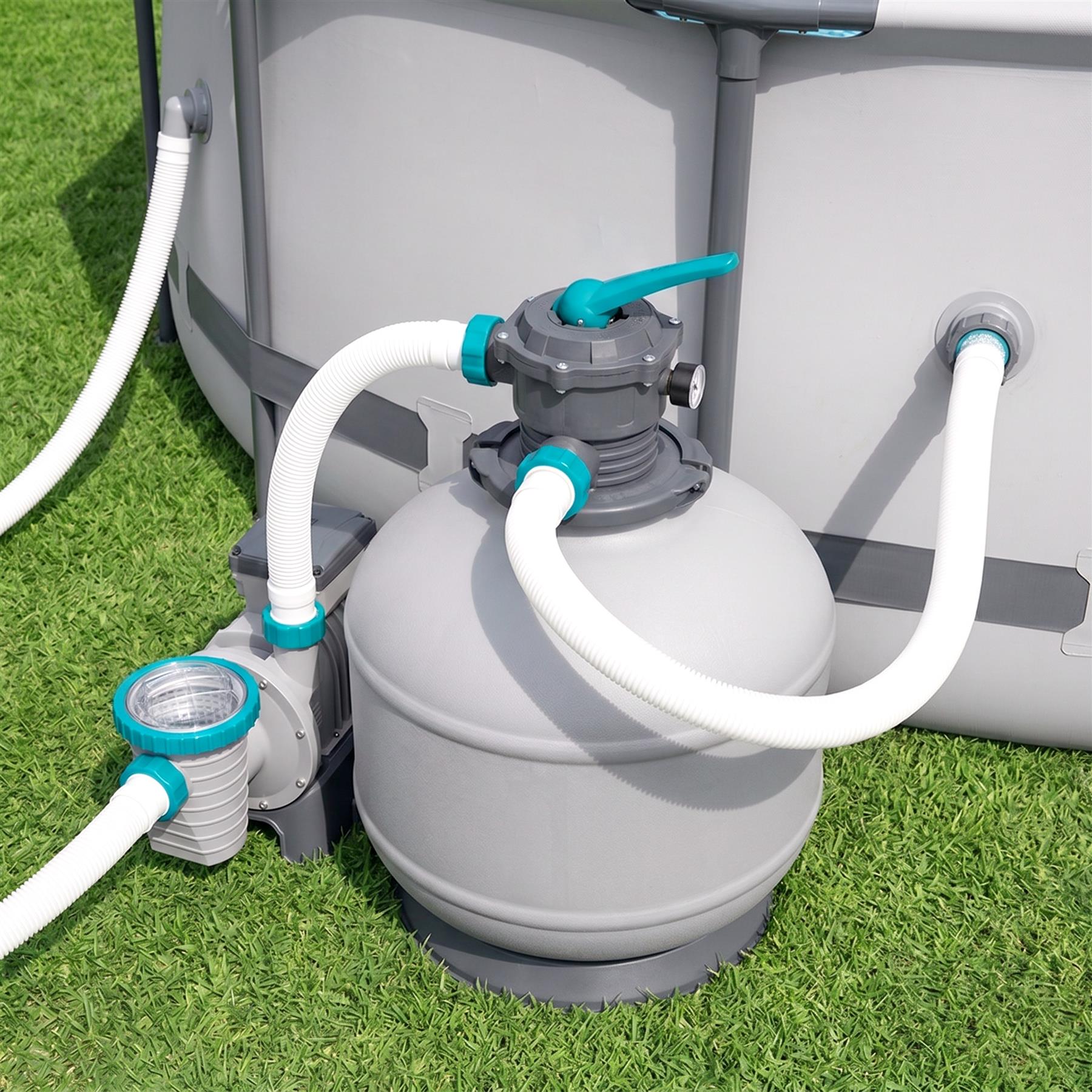 A gray filter system unit sits on green grass, connected by white hoses and blue fittings.