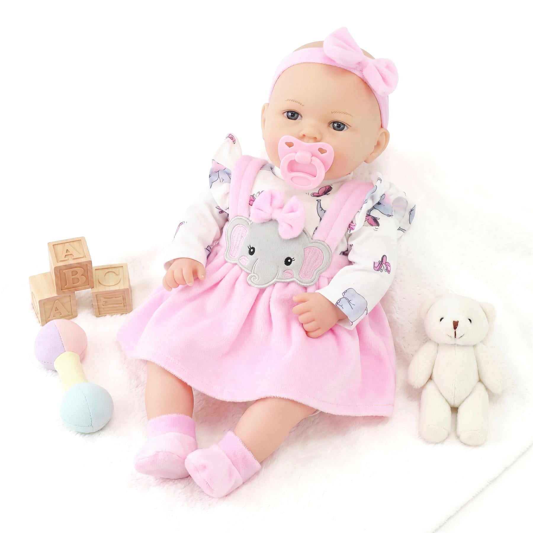 Pink baby doll surrounded by wooden alphabet blocks, a pastel rattle, and a stuffed teddy bear.