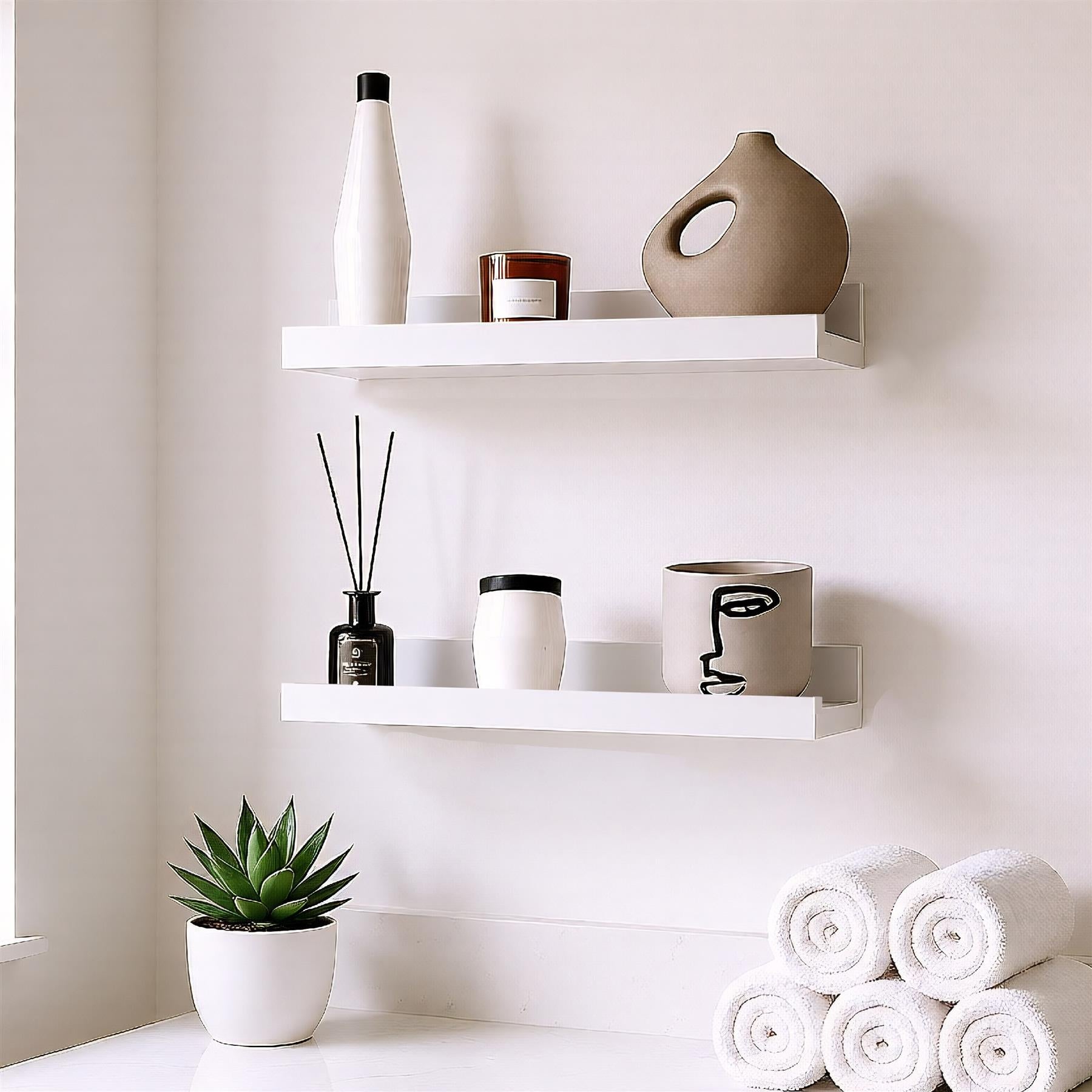 A white shelf with two shelves, holding various objects. The top shelf has a white bottle, a brown candle, and a beige vase. The bottom shelf has a black reed diffuser, a white candle, and a white pot with a black face design. Below the shelves, a small potted plant and a stack of white towels are placed. - The Magic Toy Shop