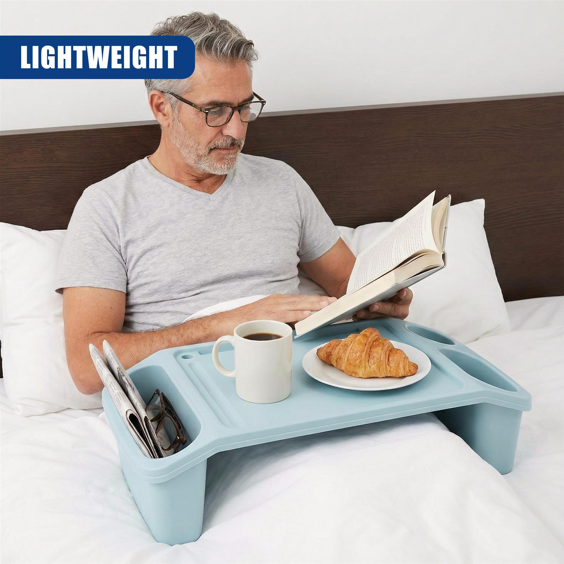 A man in bed with a tray in front of him, holding a book and eating a croissant. The tray has a cup of coffee and a plate of croissants. The man is in a comfortable position, with a pillow behind him and a blanket on his bed. The tray is a light blue color and has a few compartments for holding small items, such as a phone or a book. - The Magic Toy Shop