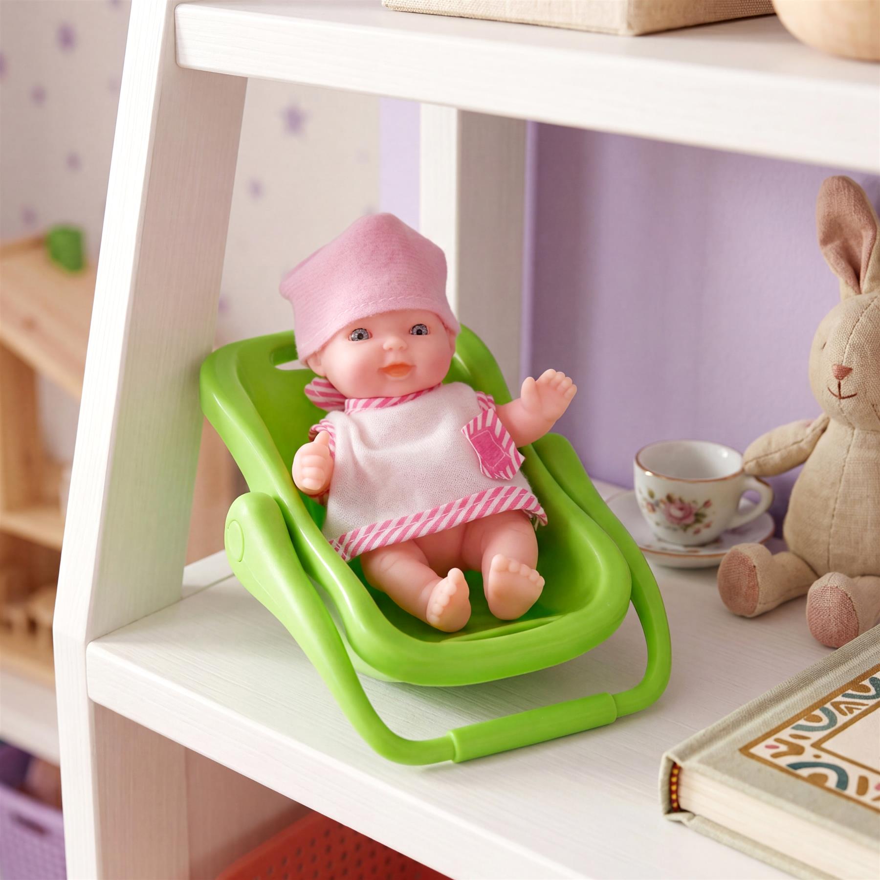 A baby doll sits in a green swing next to a stuffed rabbit and a teacup on a white shelf.