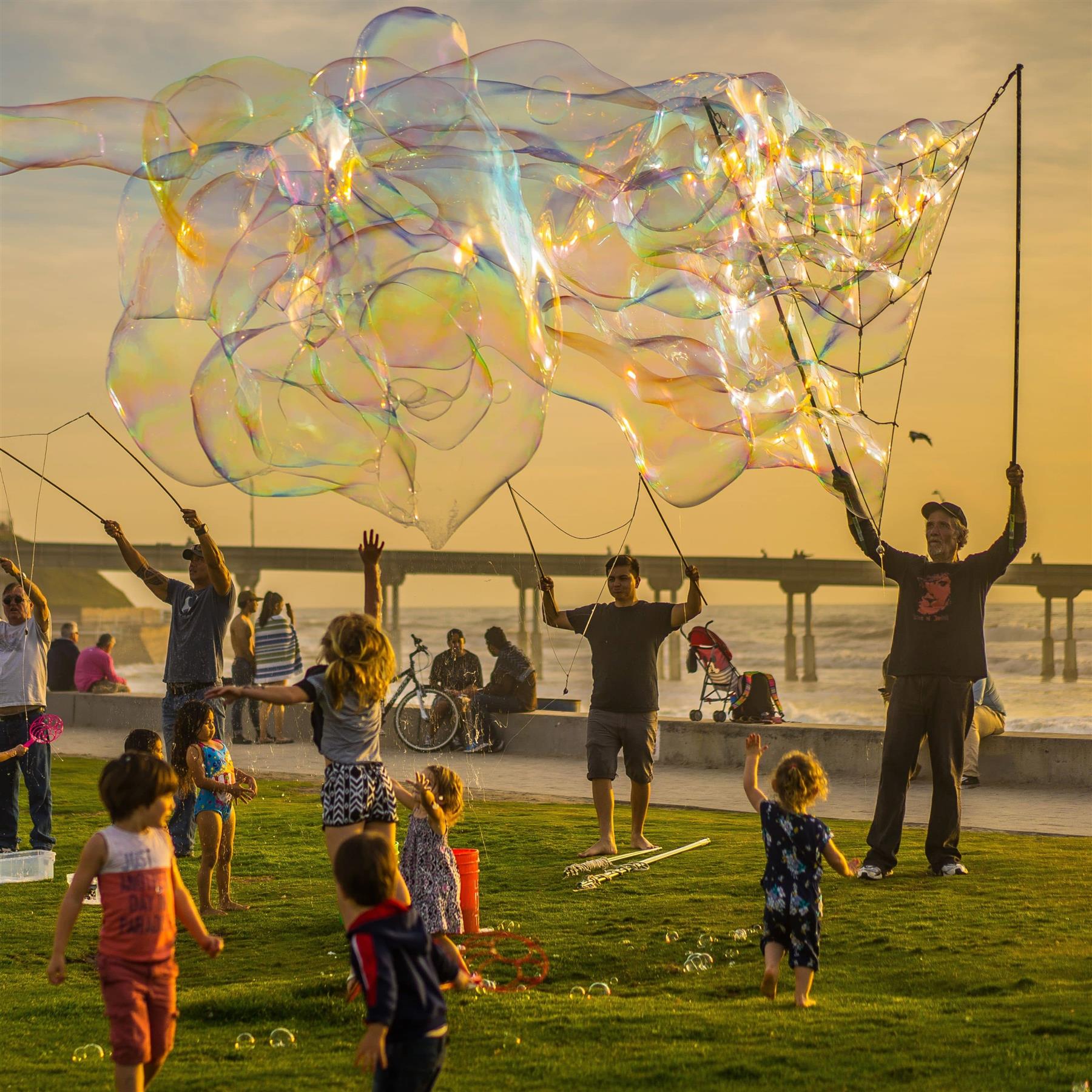 "Kids enjoying bubble play at a beach park, with large iridescent bubbles. Adults and children in various activities. People flying giant soap bubbles. Sunset by the beach." - The Magic Toy Shop