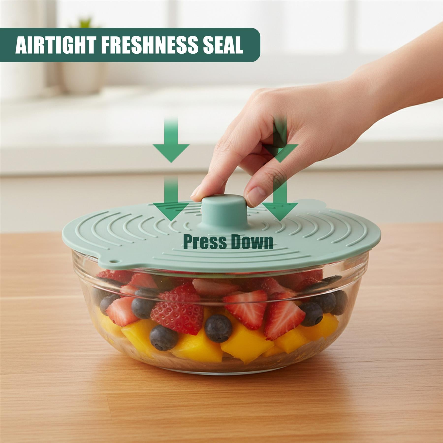 Hand presses green dome lid onto glass bowl containing sliced strawberries, mango, and blueberries.