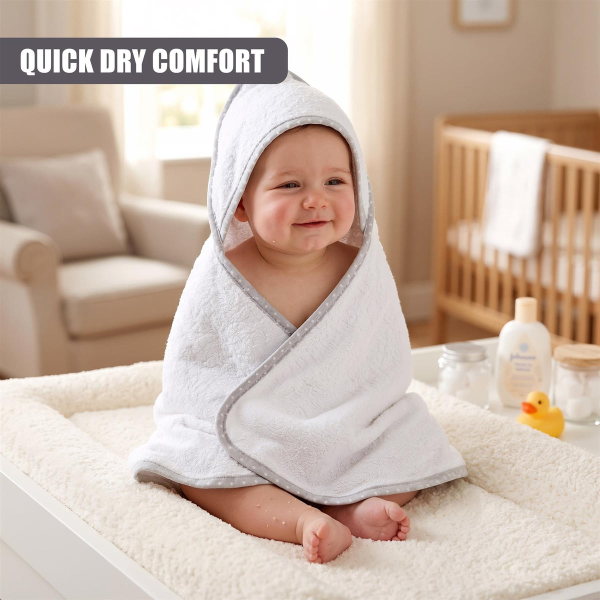 A baby wearing a grey and white hooded towel smiles in a bright nursery setting