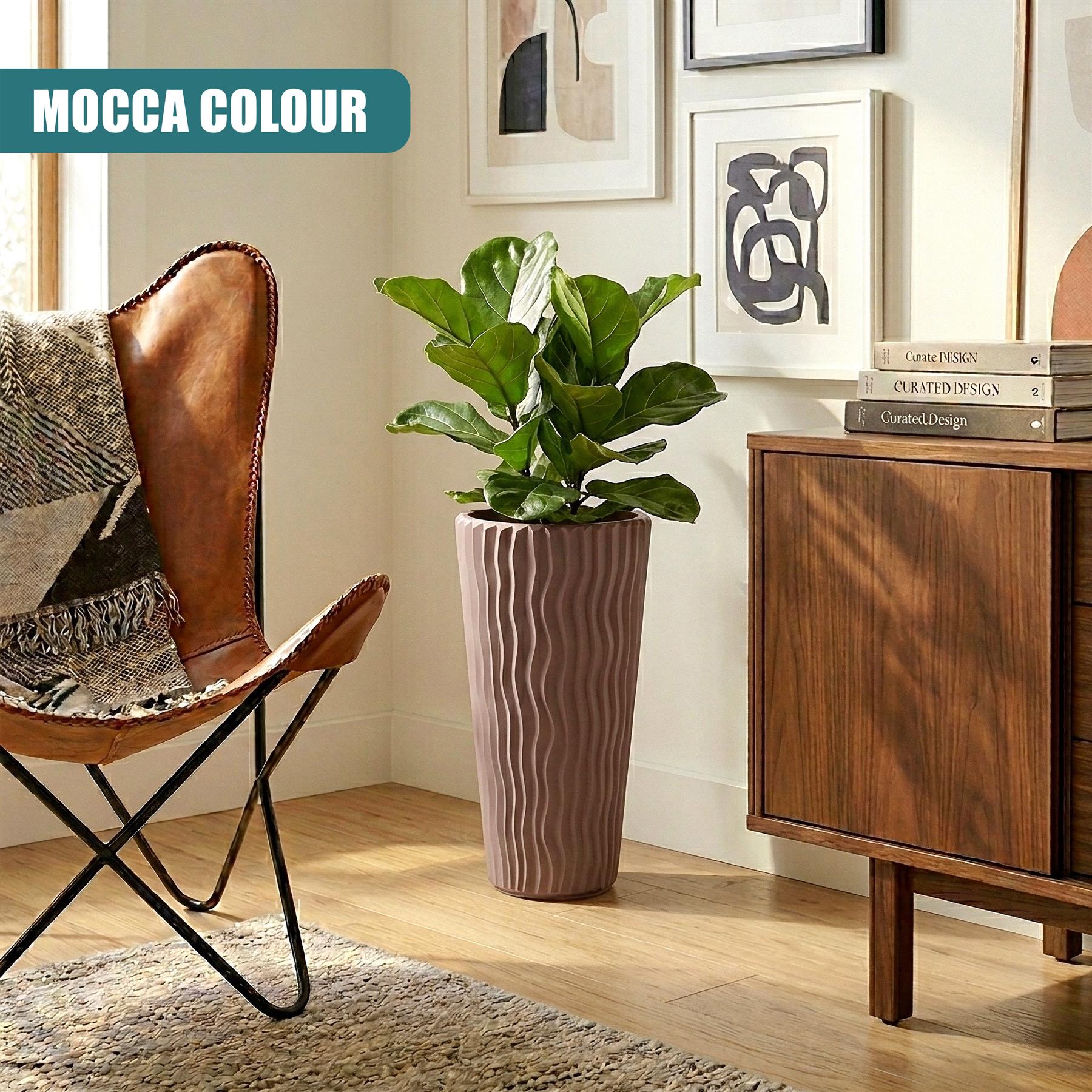 A tall, fluted, light brown planter pot holds a leafy green houseplant on a wooden floor.