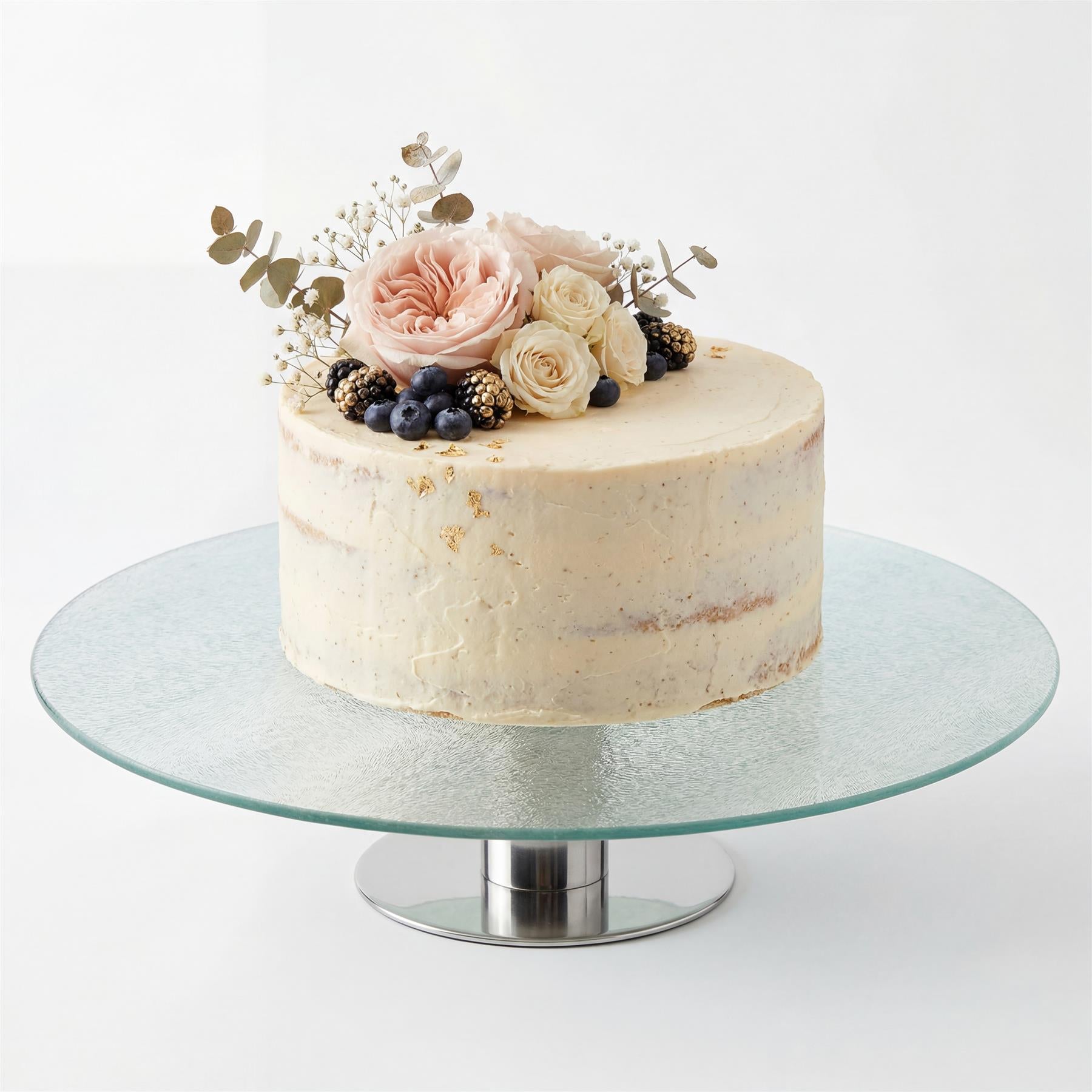 A glass cake stand displays a pale cake decorated with blue berries, pink flowers, and greenery.