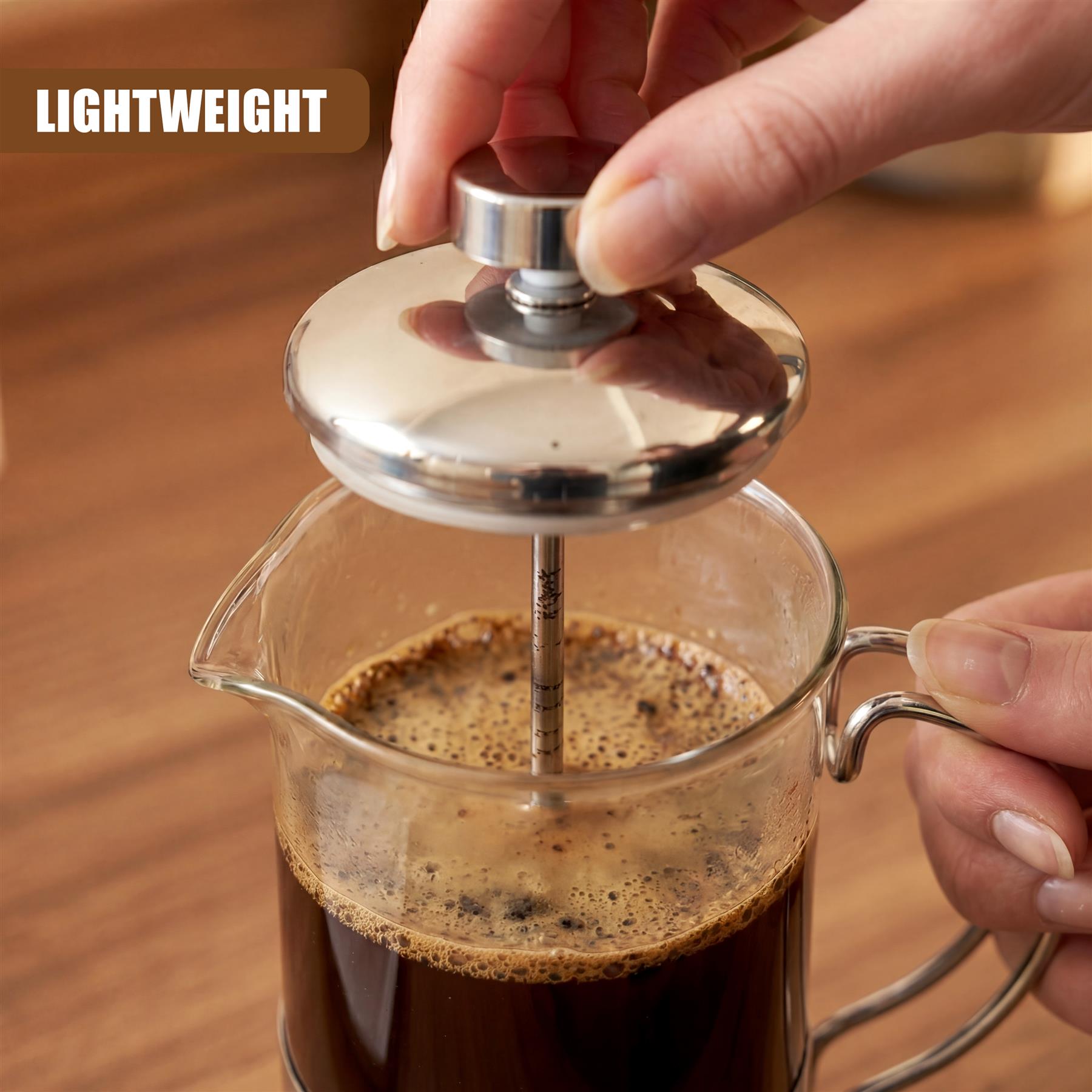 Hands use a metal plunger to press coffee through a glass carafe containing brewed coffee.