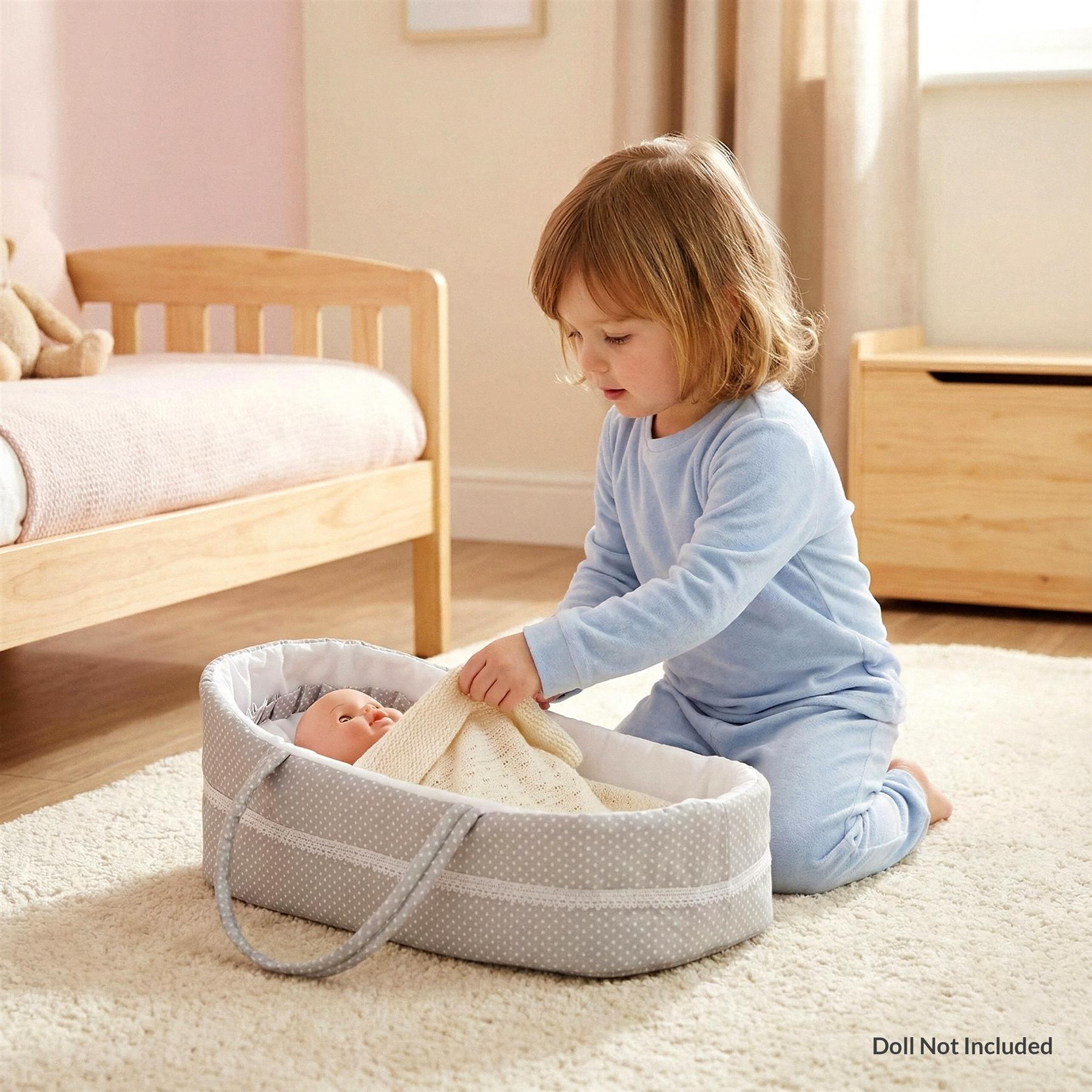 A young child playing with a baby doll in a co-sleeping bassinet, a soft and cozy bed for infants, with a built-in bassinet and storage for essentials. The child is dressed in a blue onesie and holding a soft toy, while the baby doll is wrapped in a soft blanket. The co-sleeping bassinet is placed on a soft, white carpeted floor, with a small table or desk in the background. The room is well-lit and has a calm, peaceful atmosphere. - The Magic Toy Shop