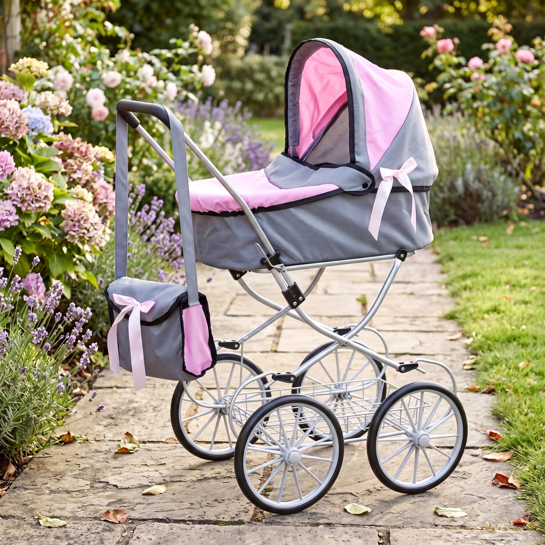 Pink and gray doll stroller displayed outdoors on a stone path surrounded by blooming flowers.