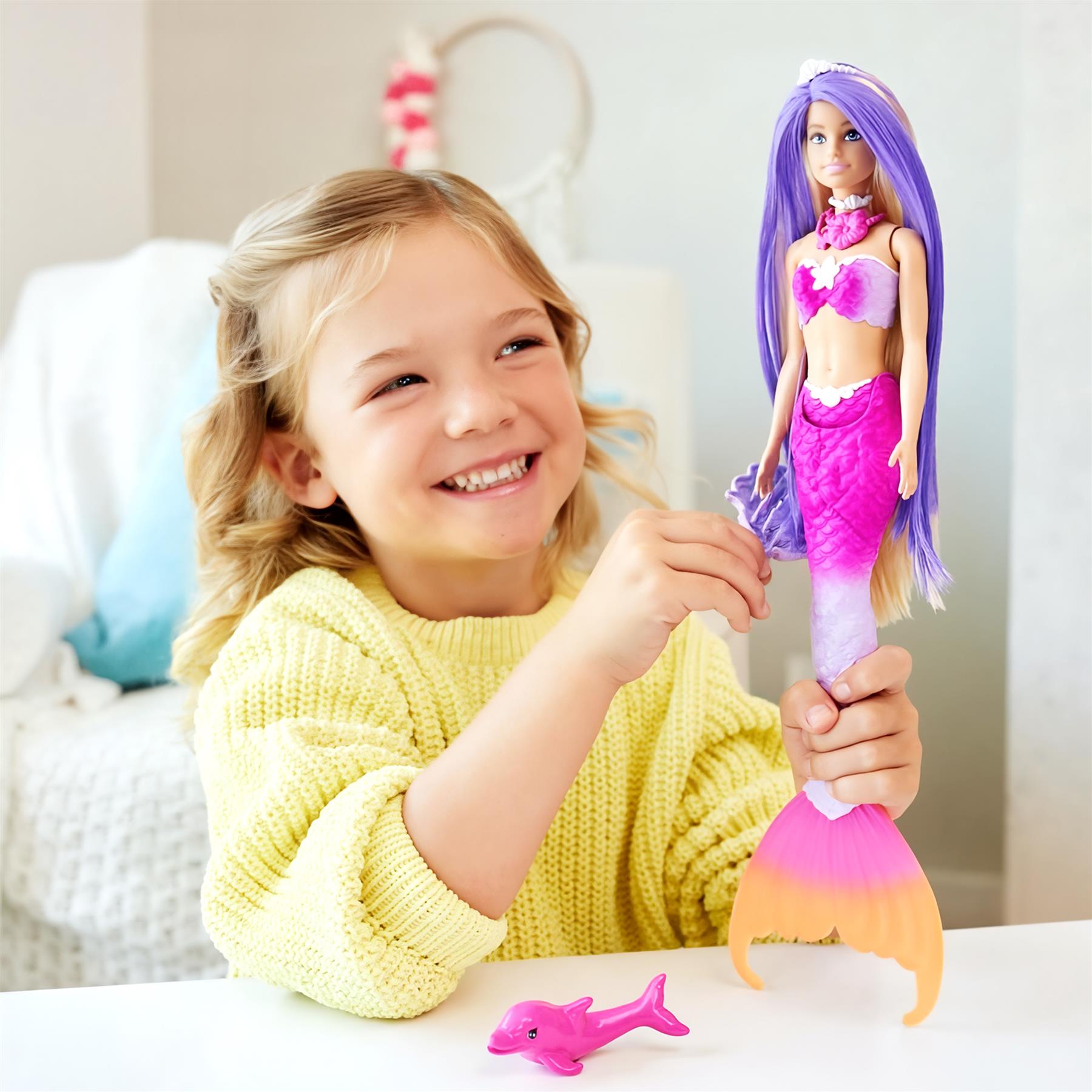 Blonde girl smiling while holding a purple-haired mermaid doll and a pink dolphin toy.