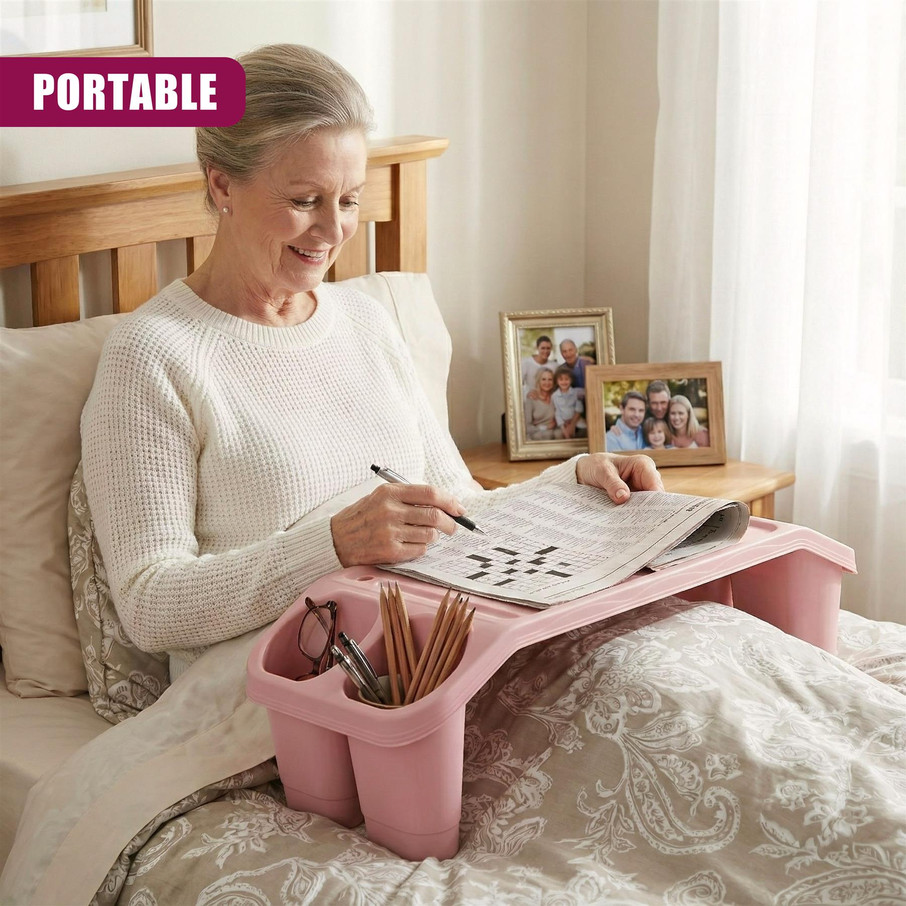 A woman sits in bed, holding a crossword puzzle and using a pink, foldable lap tray to hold her pen, glasses, and other small items. The tray has a built-in cup holder and a storage compartment. The woman is comfortable and relaxed, with a few framed family photos on her bedside table. - The Magic Toy Shop
