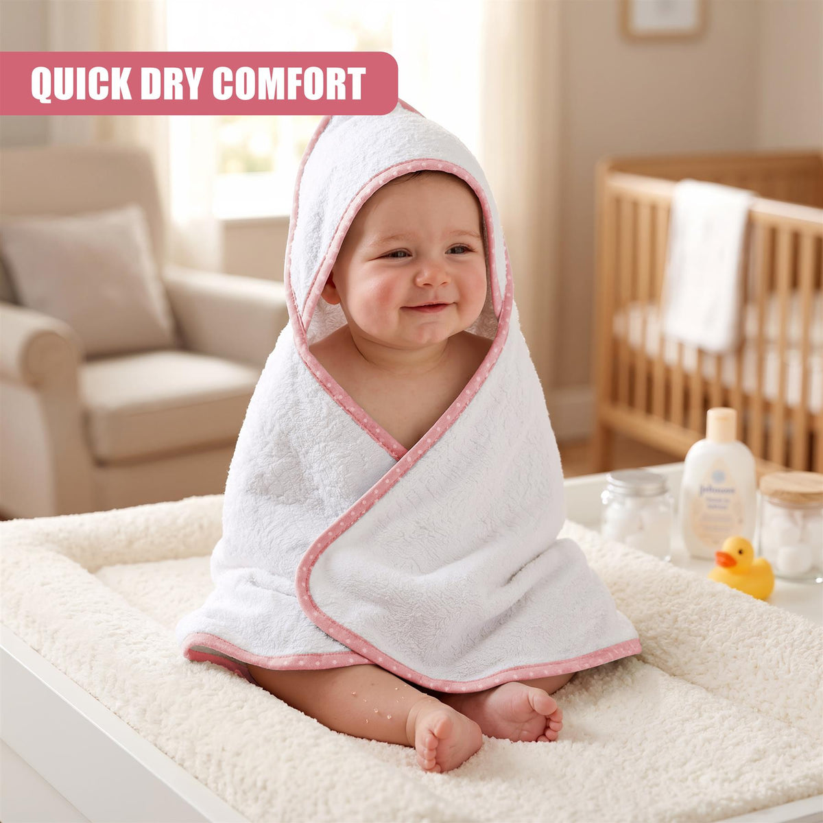 A baby wrapped in a pink and white GEEZY hooded bath towel smiles in a bright nursery setting