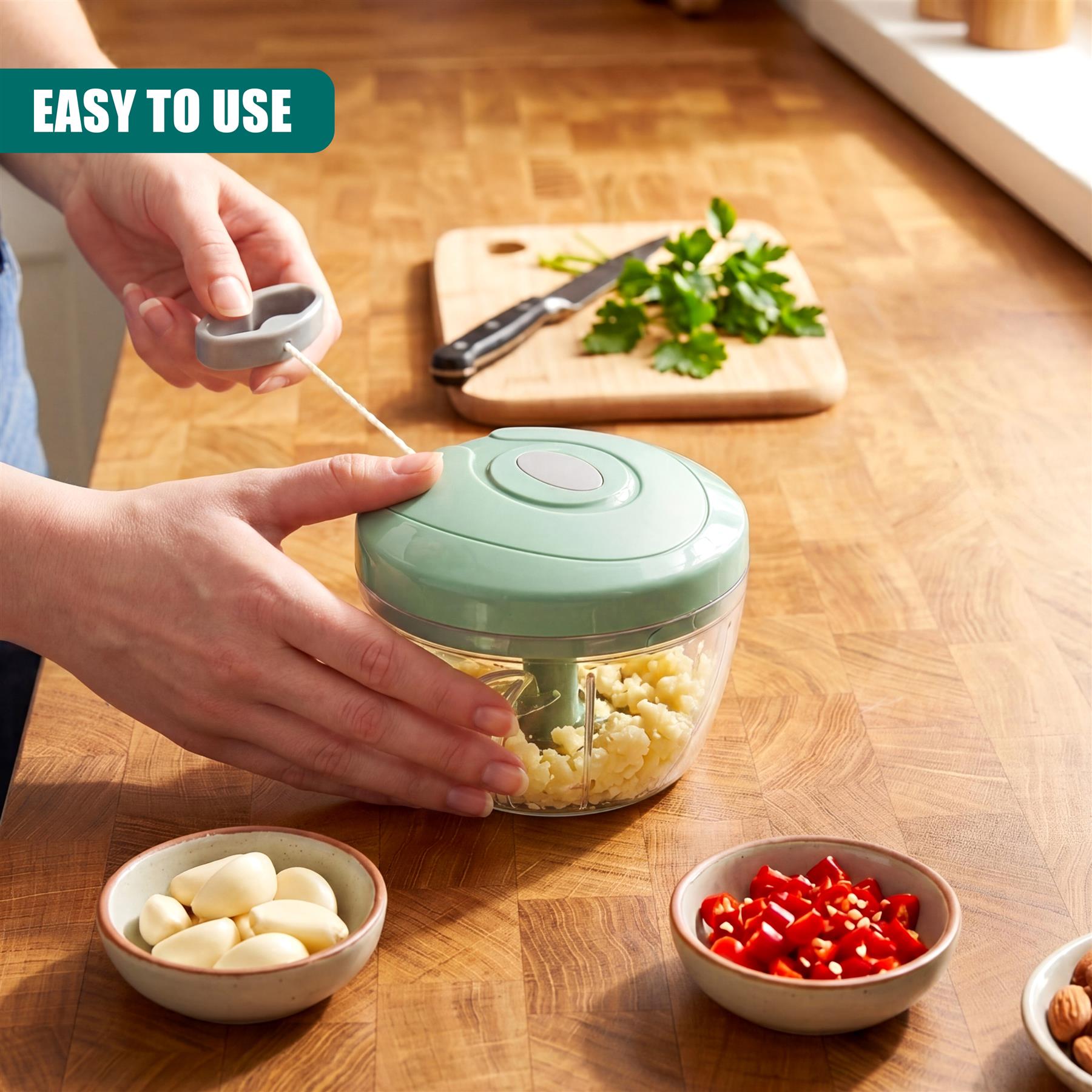 Hands operate a manual food chopper filled with herbs near bowls of garlic and chilies.