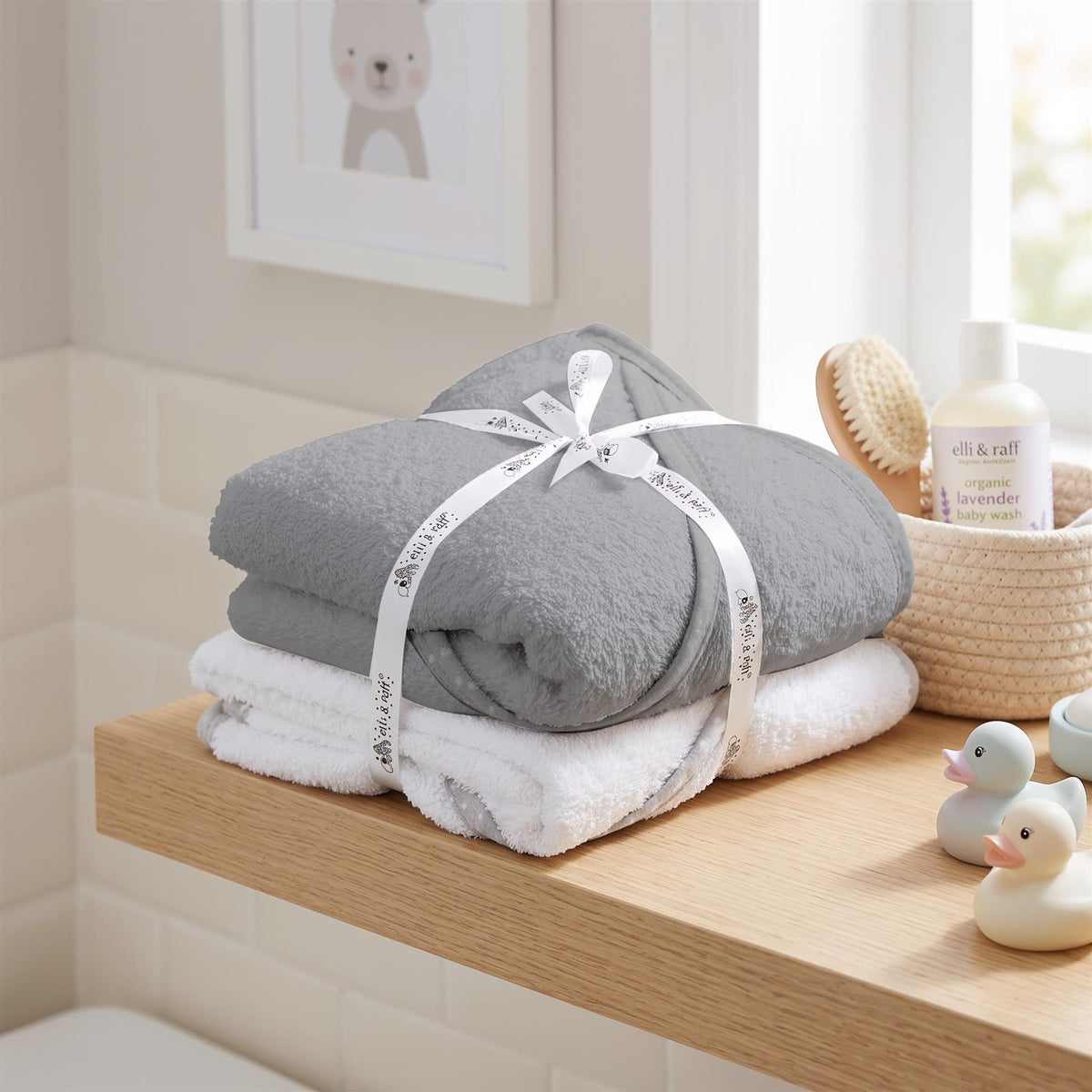 Two grey GEEZY hooded baby towels are stacked on a wooden surface with baby bath toys