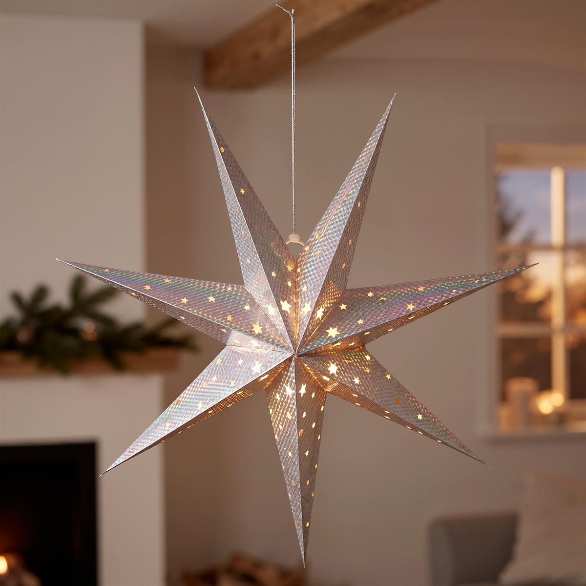 A silver LED paper star lantern hangs in a cosy room, illuminated with warm white lights