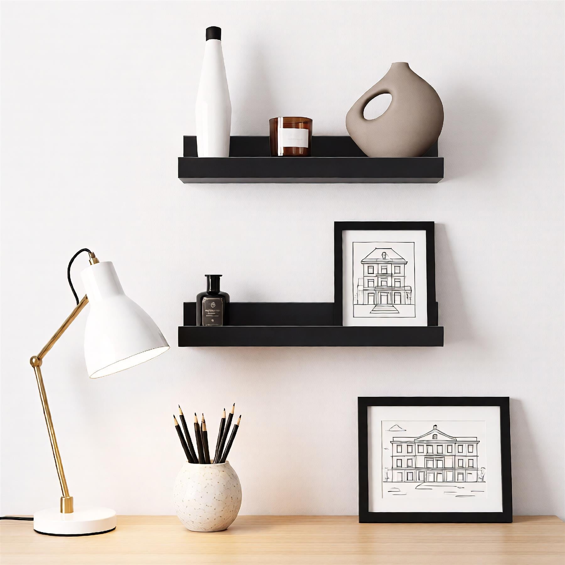 A modern shelf set-up featuring two floating shelves in a dark wood tone, each containing various decorative items. The top shelf holds a white and black bottle, a brown candle, and a beige vase. The bottom shelf features a black frame with a white and black sketch of a building, a black bottle, and a white ceramic vase. A white desk lamp with a gold arm is placed on a wooden desk, accompanied by a white ceramic vase with black pencils. - The Magic Toy Shop