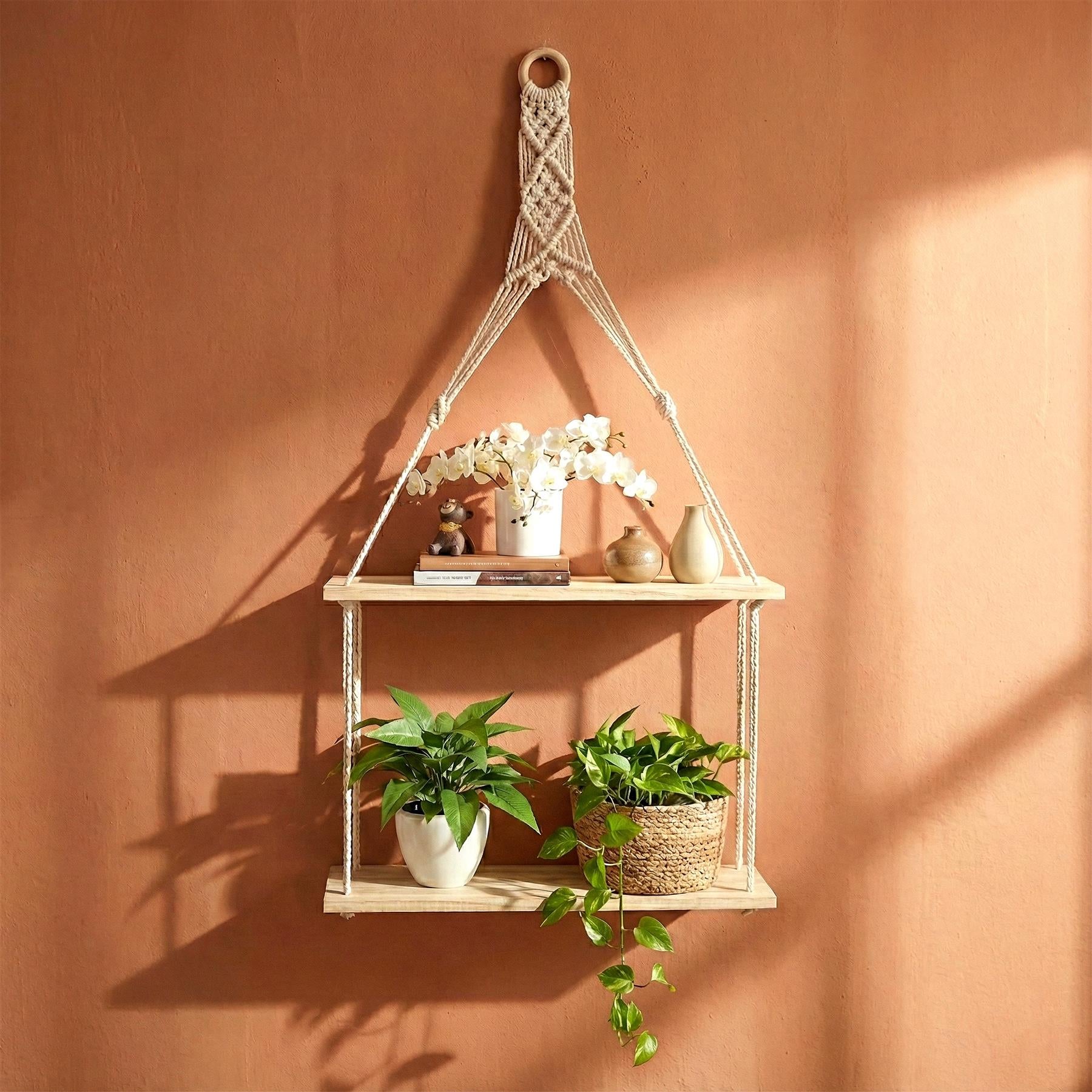 A wooden shelf with two tiers, suspended by a macramé rope, holding a plant, a book, and a few decorative items. - The Magic Toy Shop