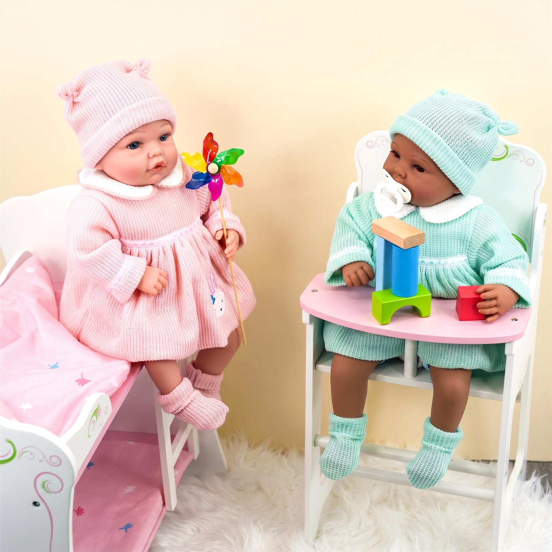 Two dolls wearing outfits—one pink, one mint green—are posed with nursery props.