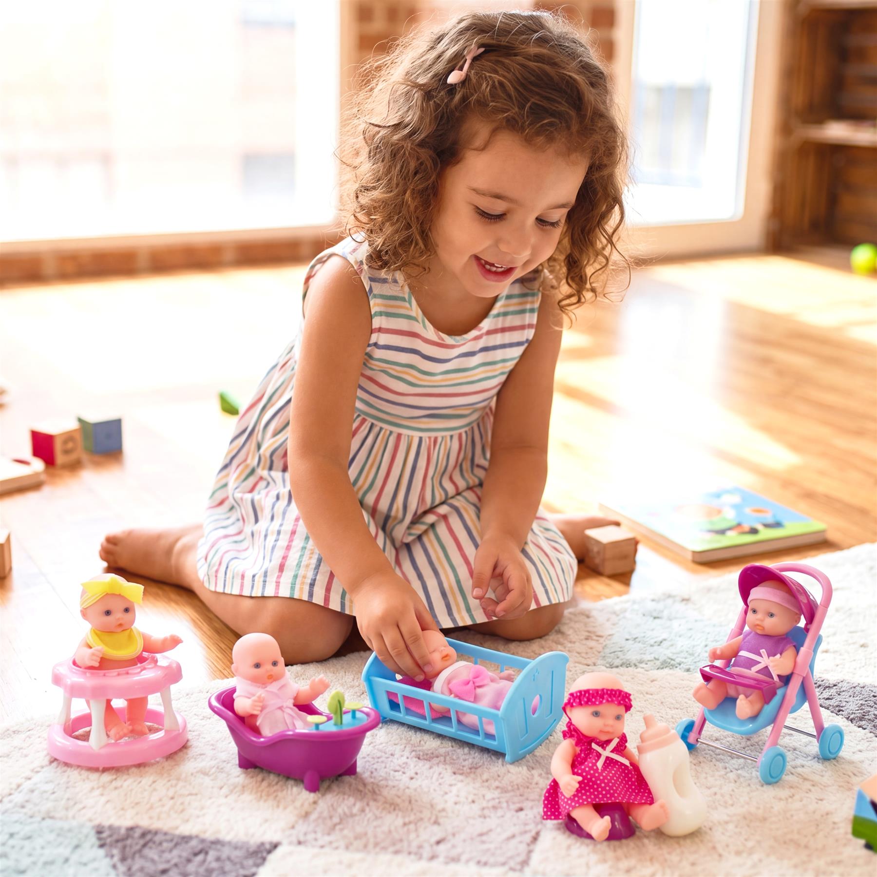A young girl plays with miniature dolls, strollers, and accessories spread across a cozy patterned rug.