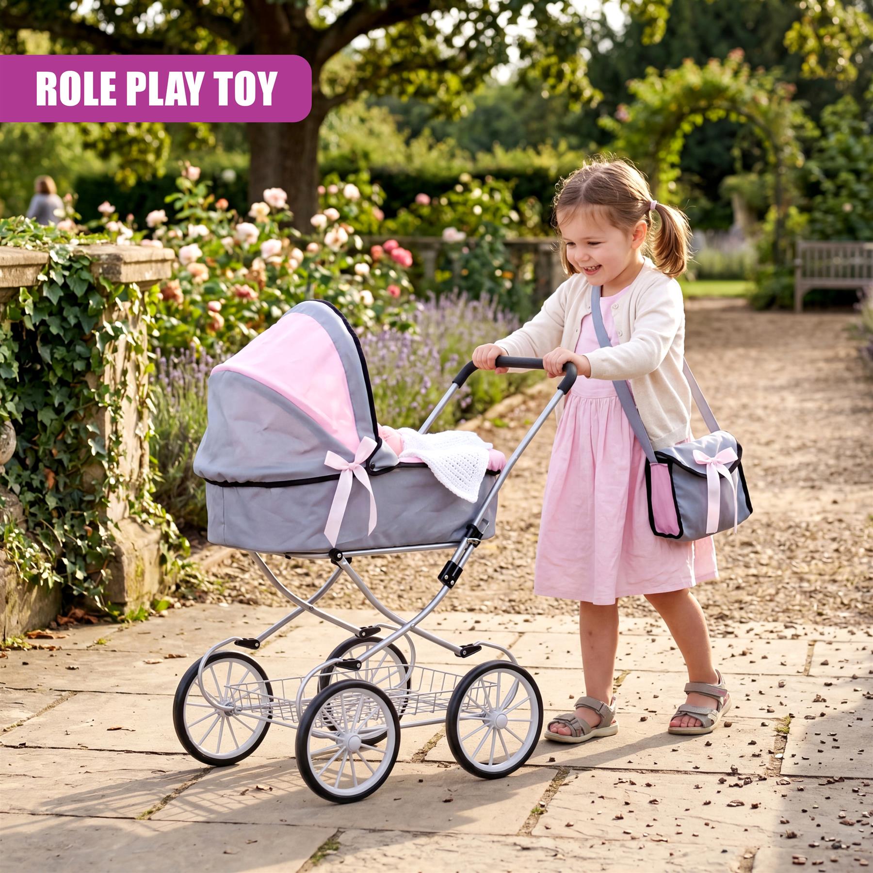 Pink and gray doll pram with wheels stands near a smiling girl in a lush outdoor garden setting.