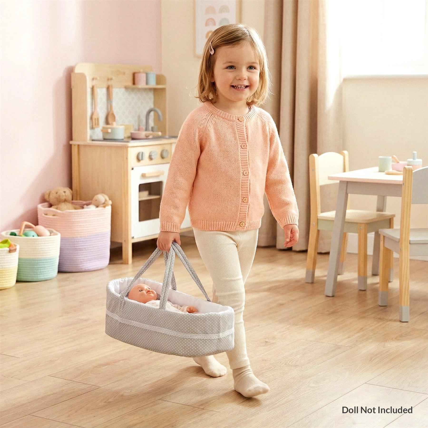 A child is holding a grey and white polka-dot baby bassinet, with a baby doll inside, in a playroom with a play kitchen, baskets, and toys. - The Magic Toy Shop