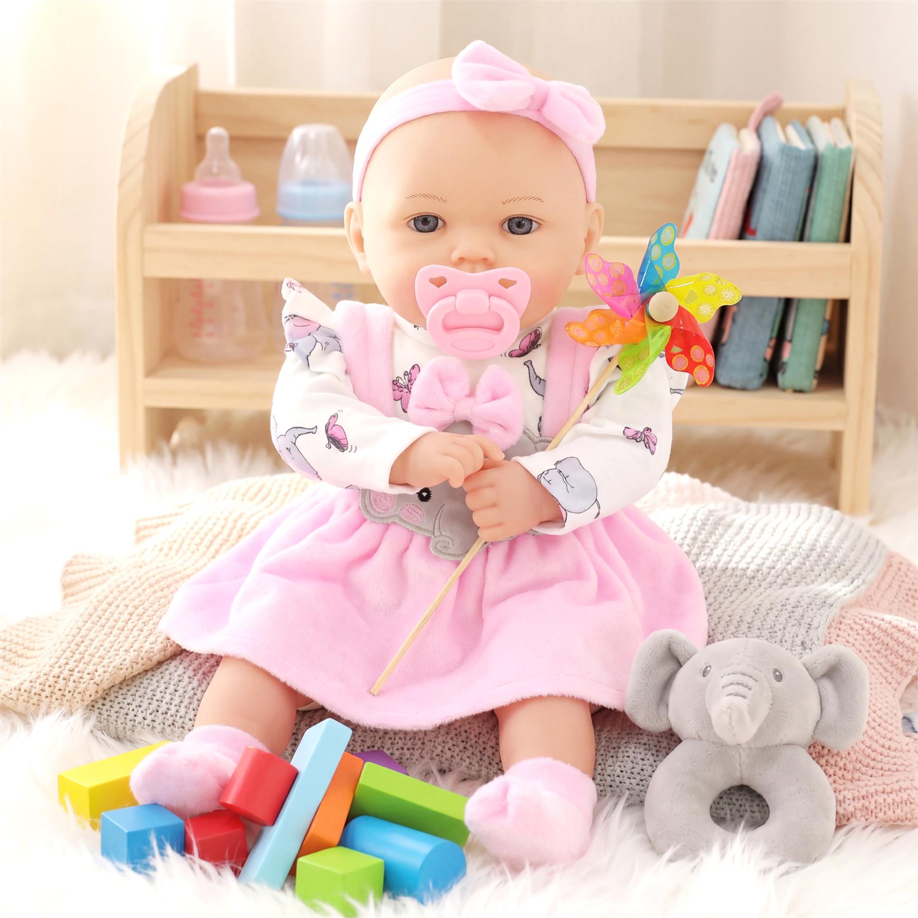 Ellie doll in a pink outfit sits with blocks and a gray stuffed elephant near a wooden crib unit.