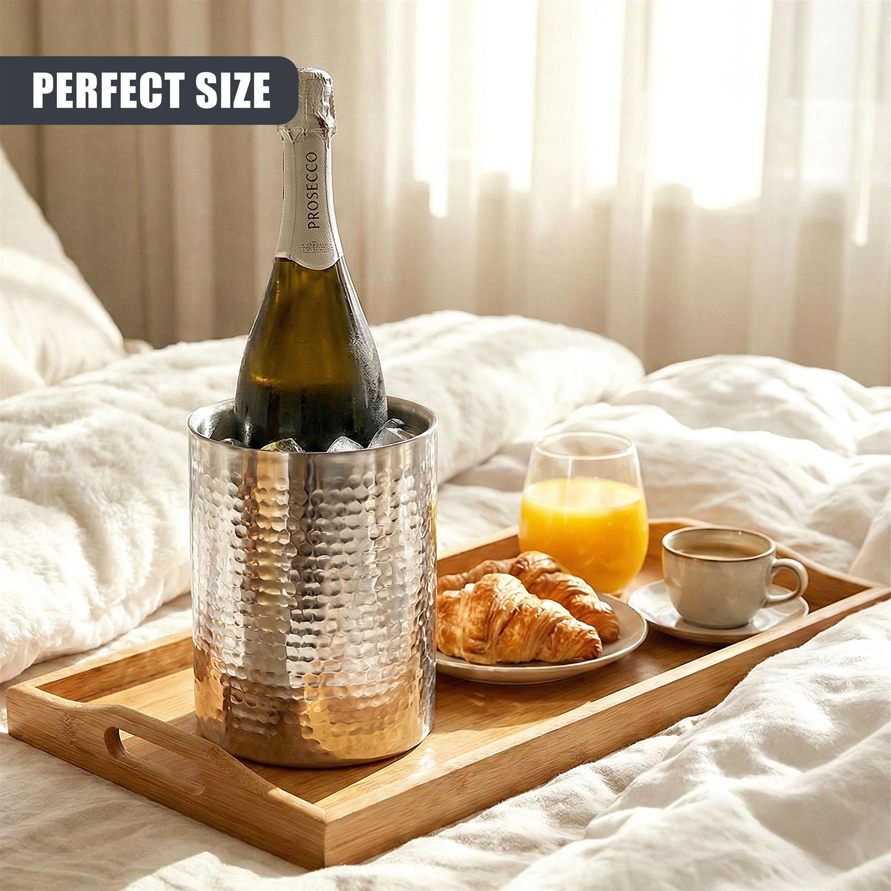 Stainless steel wine cooler on a wooden tray next to croissants, juice, and a cup of coffee.