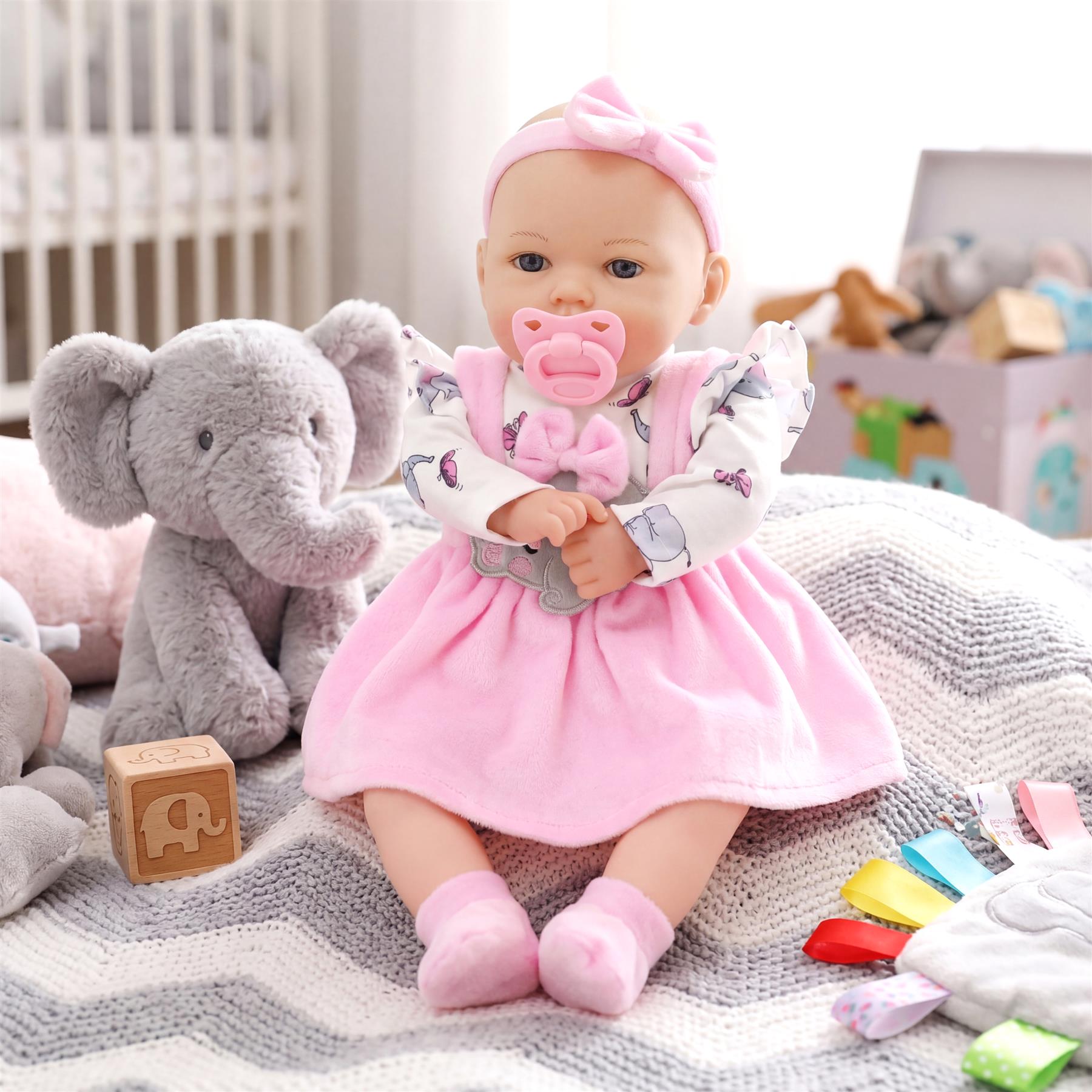 Baby doll in a pink dress sits on a patterned blanket next to a gray plush elephant toy and accessories.