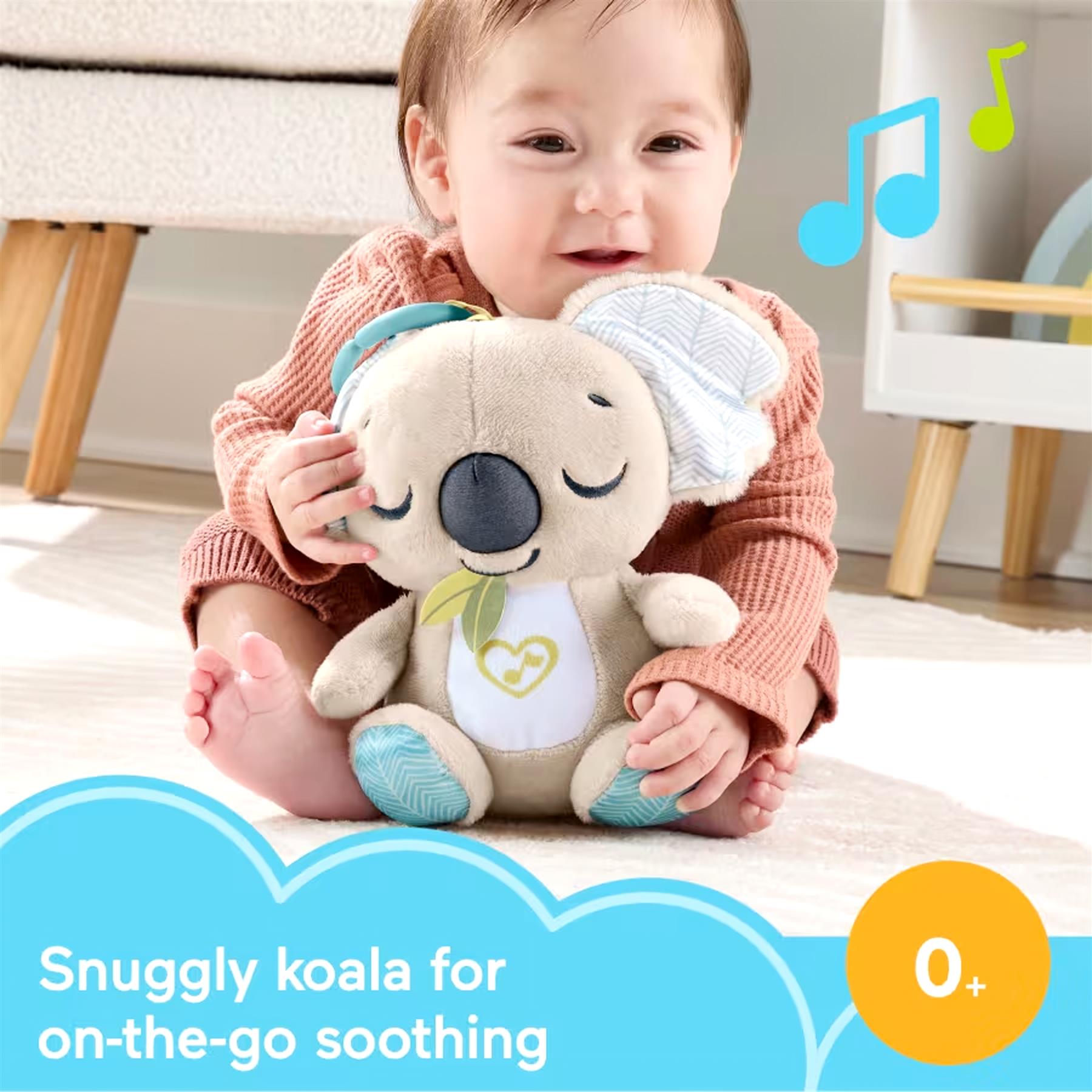 A baby sits holding a patterned plush koala toy next to a wooden dresser.
