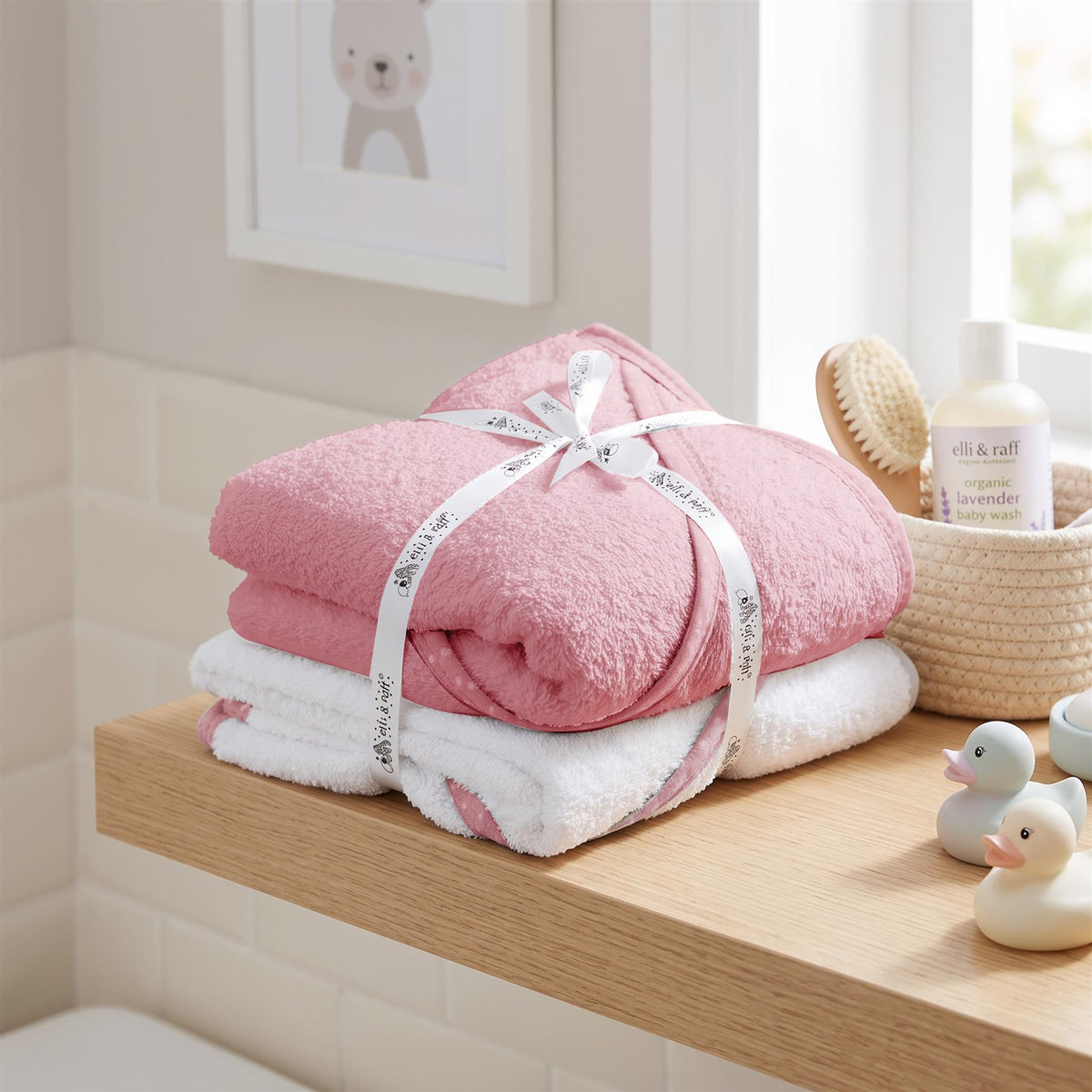 Pink hooded baby bath towels, brand GEEZY, are stacked in a bright bathroom setting