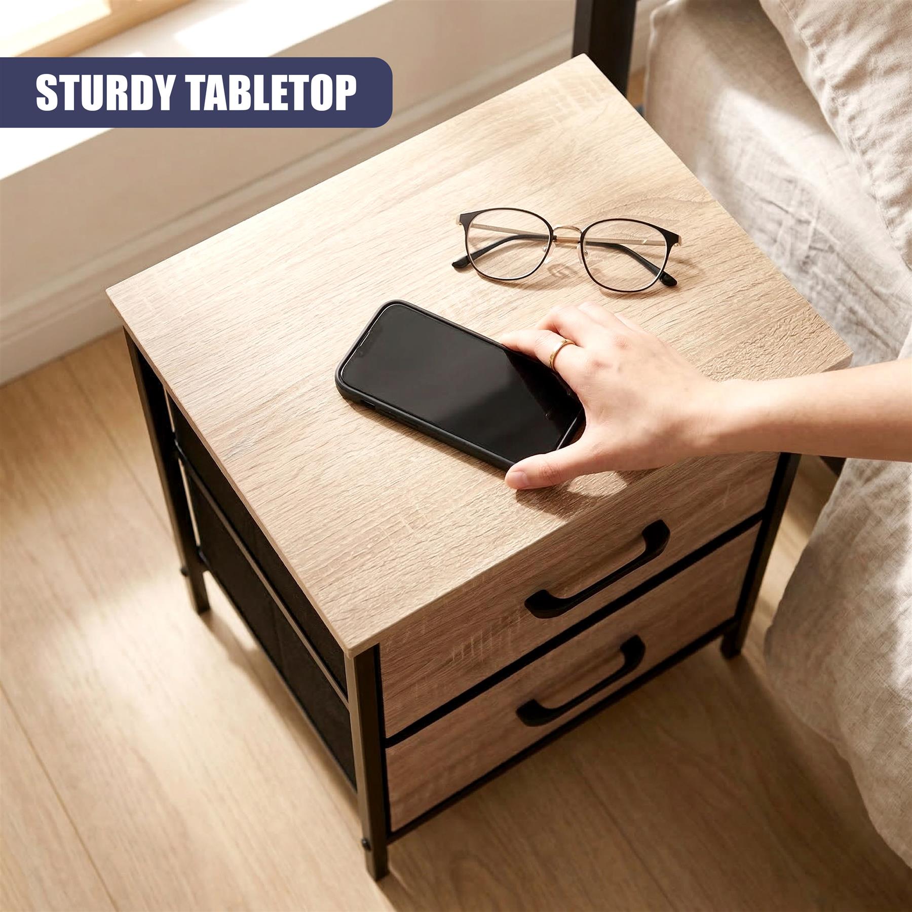 Hand resting on a light wood bedside table, displaying a smartphone, eyeglasses, and two drawers.