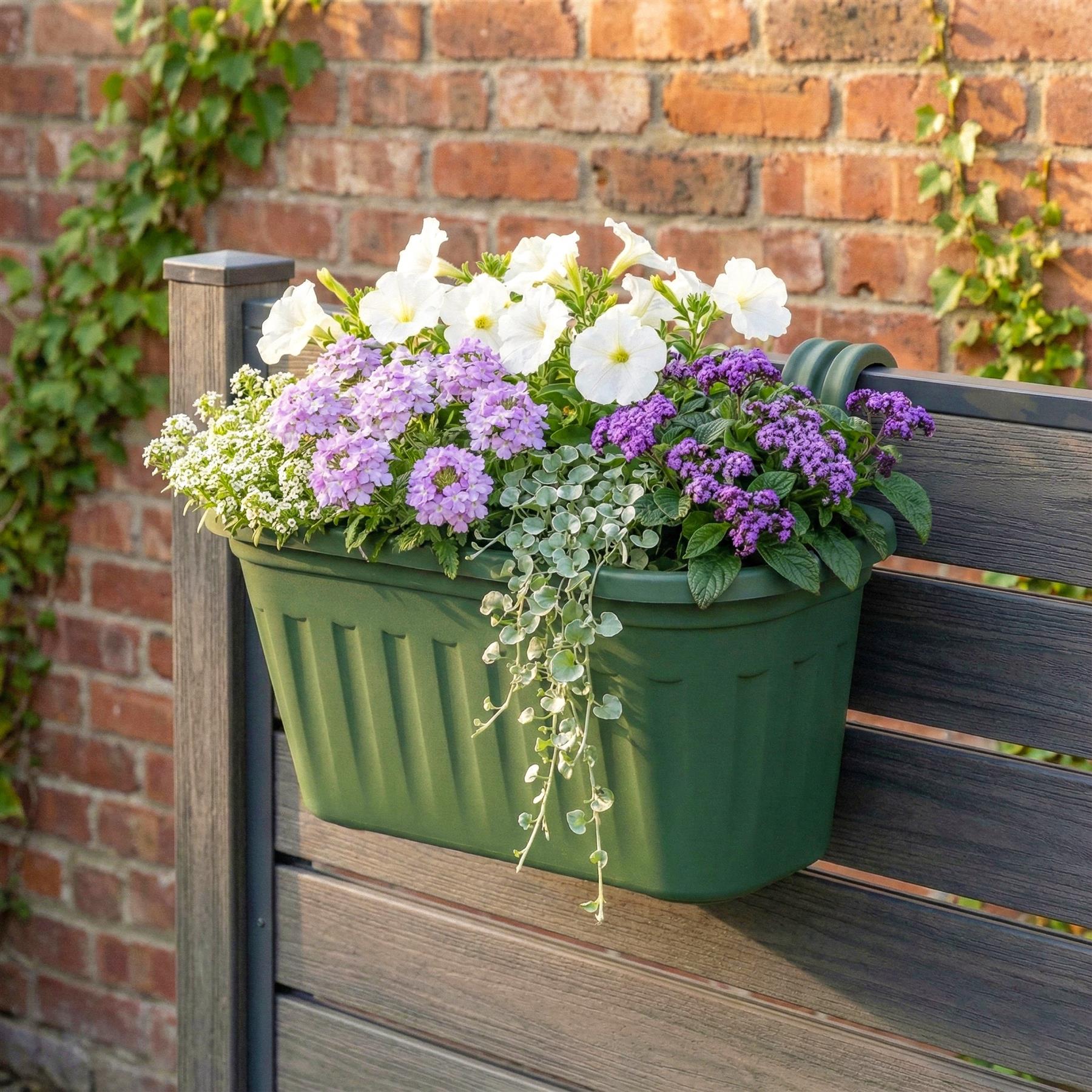 A green plastic planter with a handle, filled with a mix of white, purple, and pink flowers, and a green vine, hung on a wooden fence. - The Magic Toy Shop